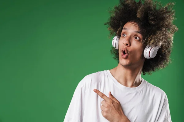 Geschokt krullend kerel poseren geïsoleerd over groene muur achtergrond luisteren muziek met koptelefoon wijzend naar copyspace. — Stockfoto