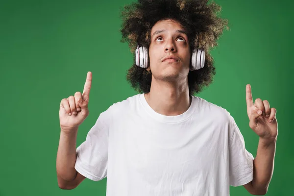 Mladý kudrnatý chlápek, který se izoloval přes zelenou zeď naslouchající hudbě se sluchátky ukazujícími na copyspace. — Stock fotografie