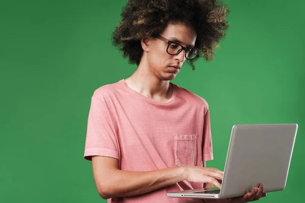 Молодой серьезный сосредоточенный кудрявый парень позирует изолированно на зеленом фоне стены с помощью ноутбука . — стоковое фото