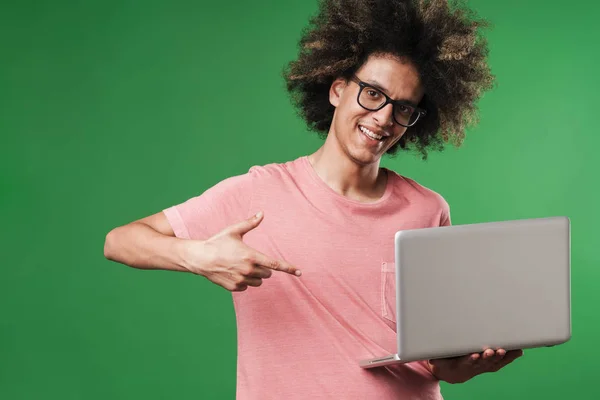 Sonriendo chico rizado feliz posando aislado sobre fondo de pared verde usando ordenador portátil . — Foto de Stock