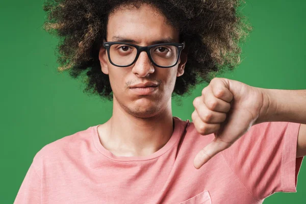 Sad displeased curly guy posing isolated over green wall background showing thumbs down. — Stock Photo, Image