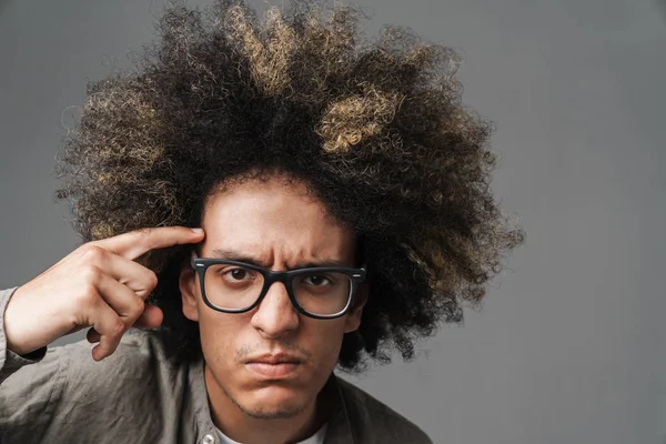 Young curly displeased teenage guy posing isolated over grey wall background showing crazy gesture holding finger near head. — Stock Photo, Image