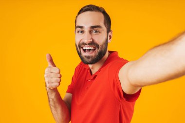 Sarı arka plan üzerinde izole selfie fotoğrafı çekerken kırmızı t-shirt gülme ve başparmak gösteren mutlu adam portresi