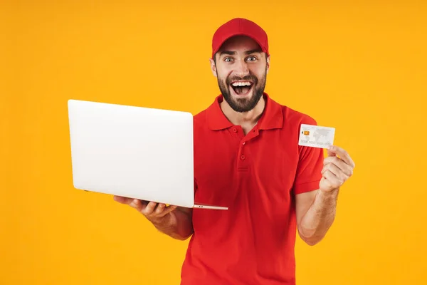 赤いTシャツとキャップを持つ楽観的な配達人の肖像画は、銀のラップトップコンピュータとクレジットカードを保持 — ストック写真