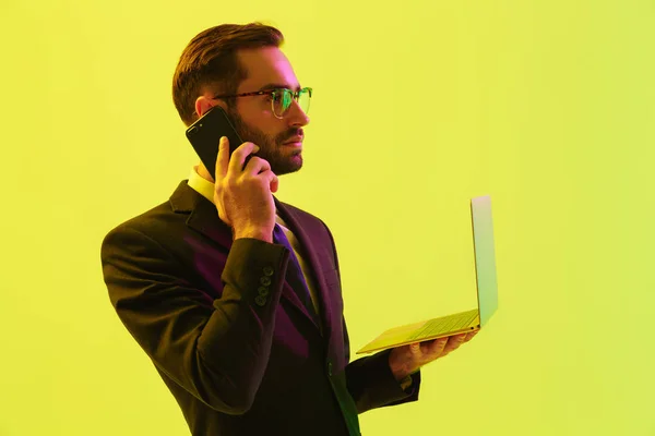 Серьезный молодой бизнесмен позирует изолированный на светло-зеленом фоне стены с ведущими неоновые огни с помощью ноутбука компьютер разговаривает по мобильному телефону . — стоковое фото