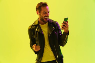Duygusal genç şık adam deri ceket giymiş led neon ışıkları ile açık yeşil arka plan duvar üzerinde izole cep telefonu ile oyun oynamak.