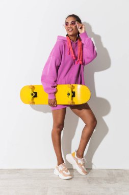 Gülümseyen memnun genç Afrikalı kadın parlak pembe sweatshirt kaykay tutan beyaz duvar arka plan üzerinde izole poz.