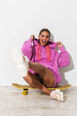 Neşeli gülümseyen iyimser genç afrikalı kadın parlak pembe sweatshirt beyaz duvar arka plan üzerinde izole kaykay üzerinde oturup.