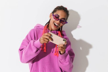 İyimser mutlu genç afrikalı kadın güneş gözlüğü giyen parlak pembe sweatshirt beyaz duvar arka plan üzerinde izole cep telefonu ile oyun oynamak.