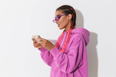 İyimser mutlu genç afrikalı kadın güneş gözlüğü giyen parlak pembe sweatshirt beyaz duvar arka plan üzerinde izole cep telefonu ile oyun oynamak.