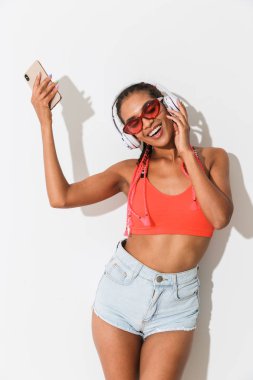 Duygusal olumlu gülümseyen genç Afrikalı kadın beyaz duvar arka plan üzerinde izole poz kulaklık dans ile cep telefonu dinleme müzik kullanarak güneş gözlüğü giyen.