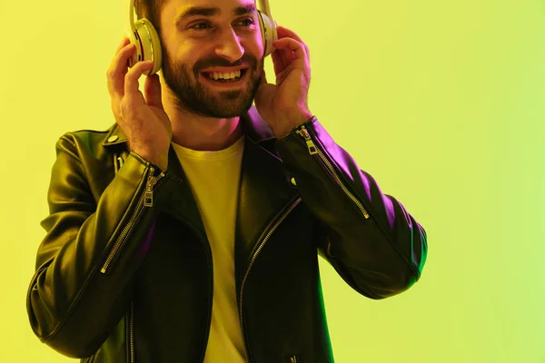 Εύθυμος ευτυχισμένος όμορφος νέος κομψός άνθρωπος που θέτει απομονωμένοι πάνω από το ανοιχτό πράσινο τοίχο φόντο με LED φώτα νέον ντυμένοι σε δερμάτινο μπουφάν ακούγοντας μουσική με ακουστικά. — Φωτογραφία Αρχείου