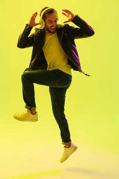 Emocional joven guapo elegante saltando bailando aislado sobre la pared de fondo verde claro con luces de neón led vestidas con chaqueta de cuero escuchando música con auriculares . — Foto de Stock