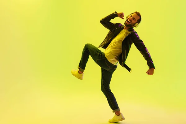 Emocional joven guapo elegante saltando bailando aislado sobre la pared de fondo verde claro con luces de neón led vestidas con chaqueta de cuero escuchando música con auriculares . —  Fotos de Stock