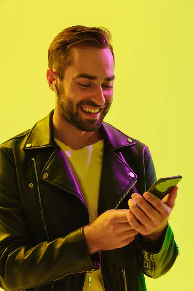 Happy cheerful handsome young stylish man isolated over light green background wall with led neon lights dressed in leather jacket using mobile phone.