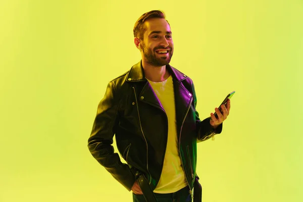 Šťastný veselý, příjemný mladý stylový muž izolovaný přes světle zelenou stěnu pozadí s neólovými světly oblečenou v koženém kabátku pomocí mobilního telefonu. — Stock fotografie