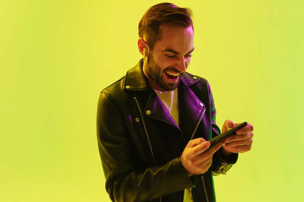 情感的年轻时尚男子隔离在浅绿色背景墙与领导霓虹灯穿着皮夹克玩手机游戏. — 图库照片