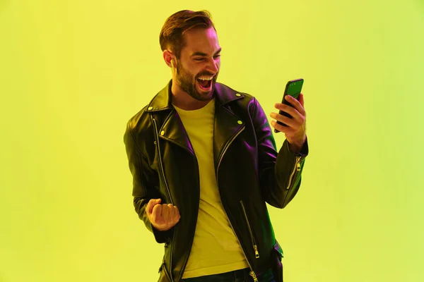 情感的年轻时尚男子隔离在浅绿色背景墙与领导霓虹灯穿着皮夹克玩手机游戏. — 图库照片
