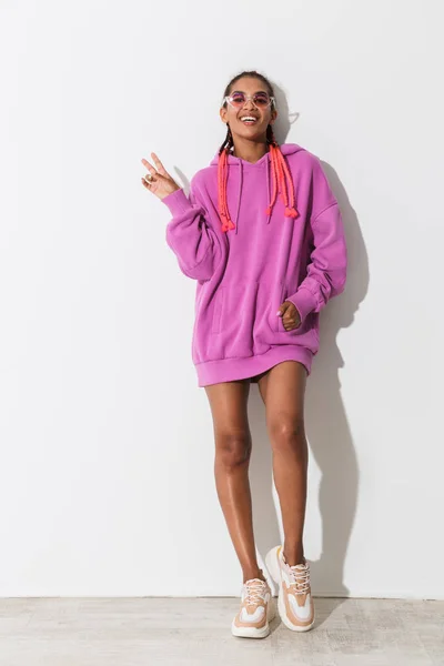 Positieve optimistische gelukkig jonge Afrikaanse stijlvolle vrouw in roze Sweatshirt en zonnebril poseren geïsoleerd over witte muur achtergrond toont vrede gebaar. — Stockfoto