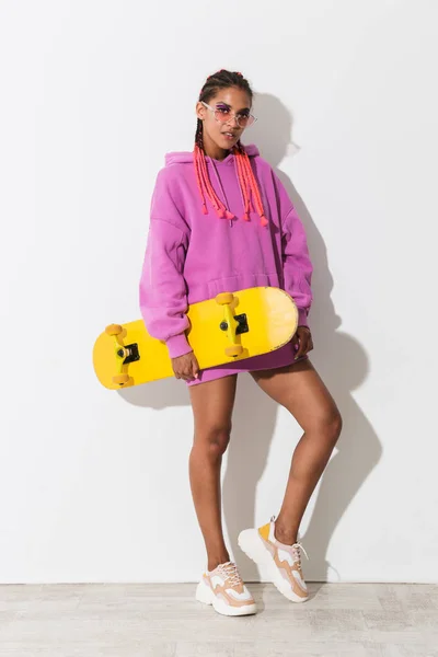 Mulher africana jovem incrível posando isolado sobre fundo da parede branca em camisola rosa brilhante segurando skate . — Fotografia de Stock