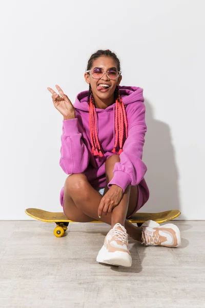 Fröhlich lächelnd optimistisch junge afrikanische Frau isoliert über weißen Wandhintergrund in leuchtend rosa Sweatshirt auf Skateboard sitzen. — Stockfoto