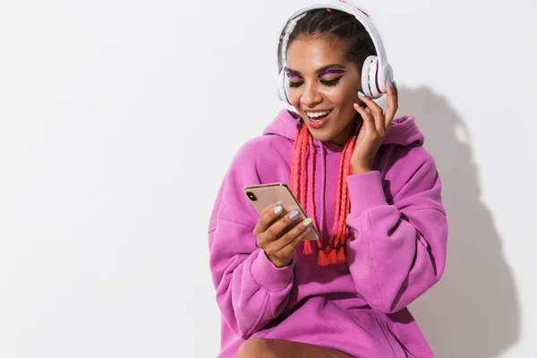 Glad positiv ung afrikansk kvinna isolerad över vit vägg bakgrund i ljust rosa sweatshirt med mobiltelefon lyssnar musik med hörlurar. — Stockfoto