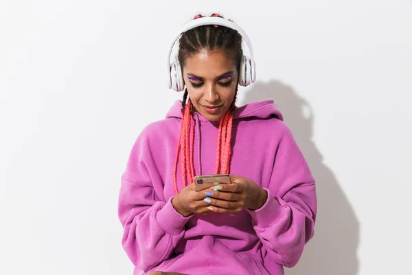 Glad positiv ung afrikansk kvinna isolerad över vit vägg bakgrund i ljust rosa sweatshirt med mobiltelefon lyssnar musik med hörlurar. — Stockfoto