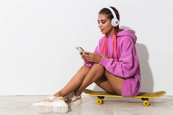 Glad ung afrikansk kvinna isolerad över vit vägg bakgrund i ljust rosa tröja sitta på skateboard med mobiltelefon lyssnar musik med hörlurar. — Stockfoto