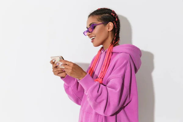 Optimistisk lycklig ung afrikansk kvinna isolerad över vit vägg bakgrund i ljust rosa tröja bär solglasögon spela spel med mobiltelefon. — Stockfoto