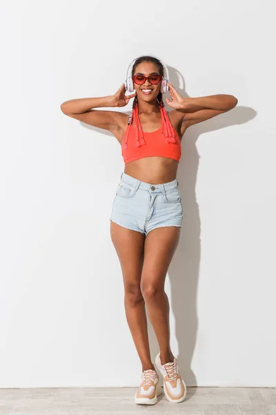Positive jeune africaine élégante femme dans des lunettes de soleil posant isolé sur fond blanc mur écouter de la musique avec écouteurs . — Photo