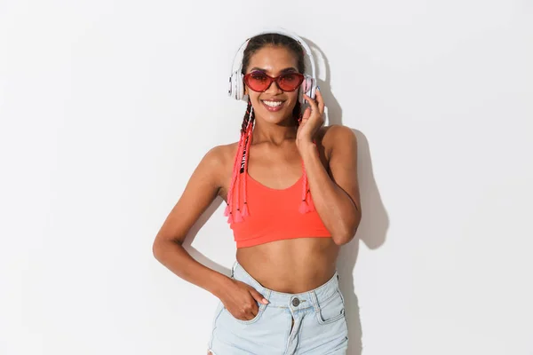 Позитивна молода африканська стильна жінка в сонцезахисних окулярах позує ізольовано на фоні білої стіни, слухаючи музику з навушниками . — стокове фото