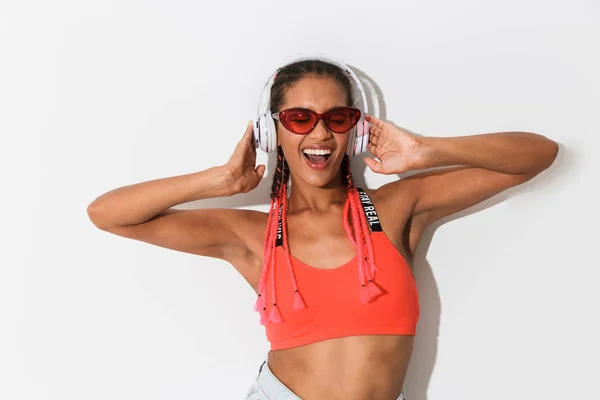 Позитивна оптимістична молода африканська стильна жінка в сонцезахисних окулярах позує ізольовано на білому фоні стіни, слухаючи музику з навушниками . — стокове фото