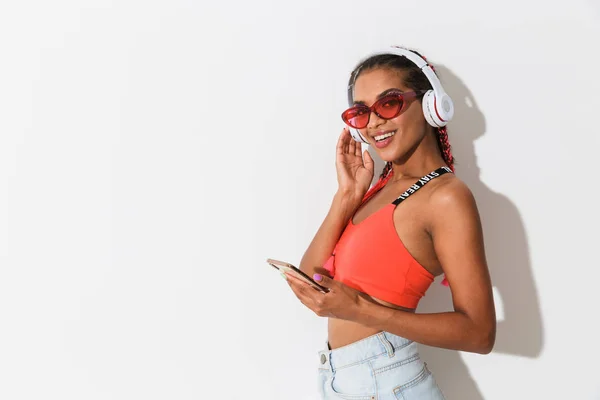 Positiv glad leende ung afrikansk kvinna poserar isolerade över vit vägg bakgrund bär solglasögon med mobiltelefon lyssnar musik med hörlurar. — Stockfoto