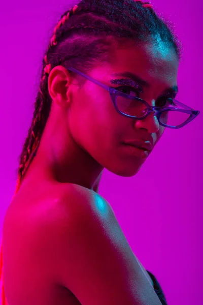 Attraente bella giovane donna africana nuda posa isolata su luci viola al neon sfondo della parete indossando occhiali da sole . — Foto Stock
