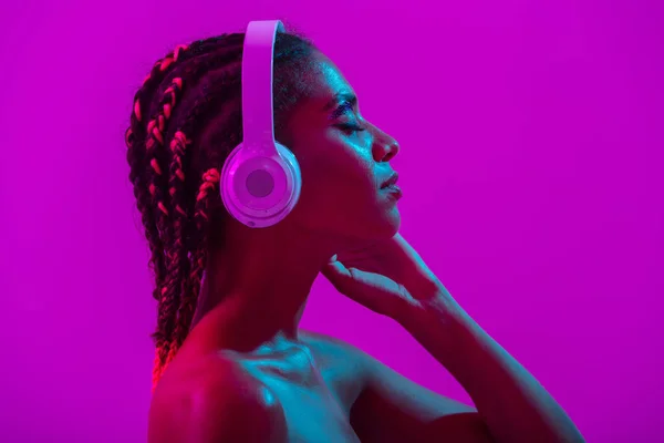 Incredibile bella giovane donna africana nuda posa isolata su luci viola al neon parete di sottofondo ascoltare musica con le cuffie . — Foto Stock