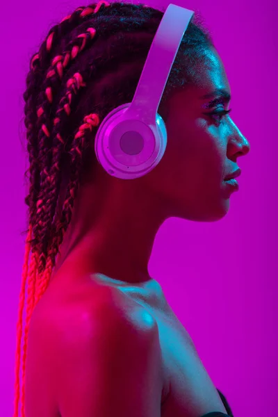 Incredibile bella giovane donna africana nuda posa isolata su luci viola al neon parete di sottofondo ascoltare musica con le cuffie . — Foto Stock