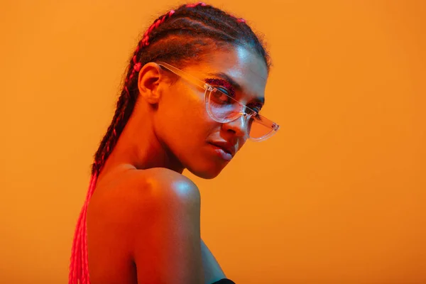 Güzel genç afrikalı çıplak kadın güneş gözlüğü giyen sarı neon ışıkları duvar arka plan üzerinde izole poz. — Stok fotoğraf