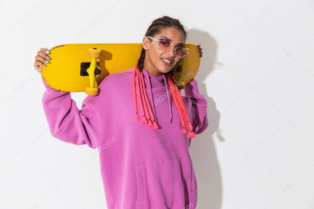 Smiling pleased young african woman posing isolated over white wall background in bright pink sweatshirt holding skateboard.