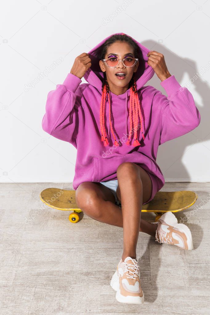 Amazing young african woman isolated over white wall background in bright pink sweatshirt sit on skateboard.