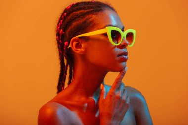 Güzel genç afrikalı çıplak kadın güneş gözlüğü giyen sarı neon ışıkları duvar arka plan üzerinde izole poz.
