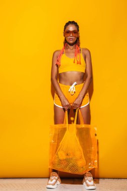 Ayakta ve plastik torba taşıyan güneş gözlüğü takan ciddi Afro-Amerikan kadın tam uzunlukta görüntü