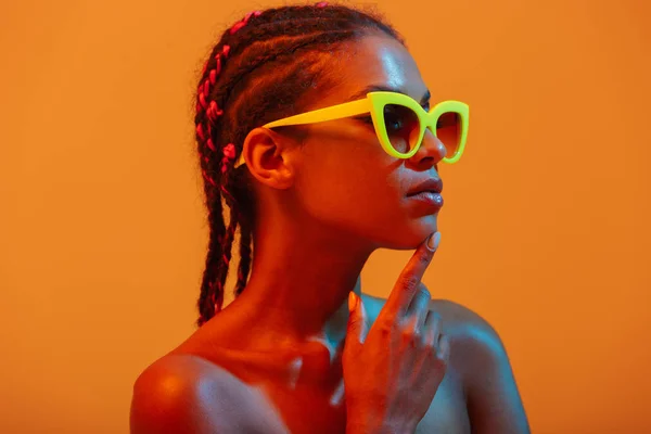Bella giovane donna africana nuda posa isolata su luci al neon gialle sfondo della parete indossando occhiali da sole . — Foto Stock