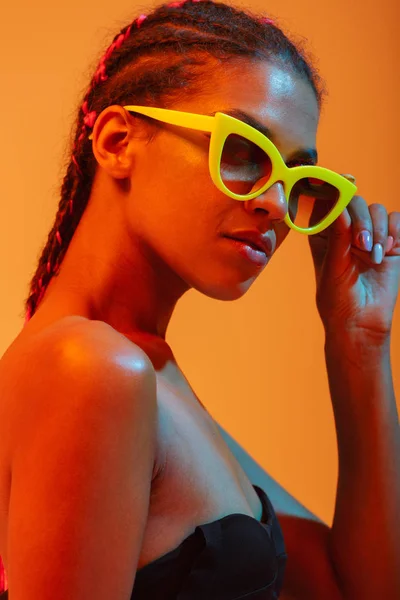Bella giovane donna africana nuda posa isolata su luci al neon gialle sfondo della parete indossando occhiali da sole . — Foto Stock