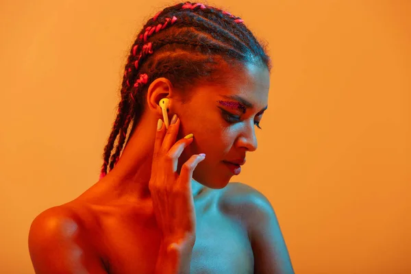 Incredibile bella giovane donna africana nuda posa isolata su luci al neon gialle sfondo della parete ascoltare musica con gli auricolari . — Foto Stock