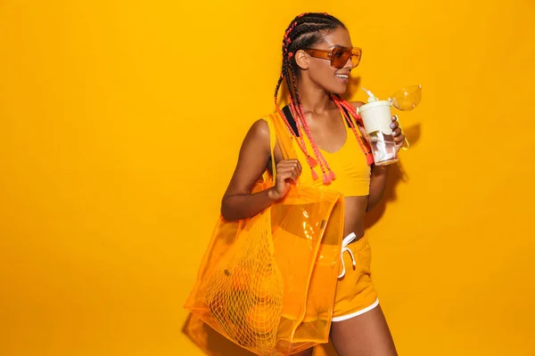 Immagine di felice donna afro-americana che indossa occhiali da sole portando borsa e tenendo tazza di plastica — Foto Stock