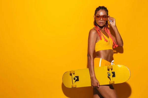 Изображение серьезной африканской женщины в солнцезащитных очках, держащей скейтборд и смотрящей в сторону — стоковое фото