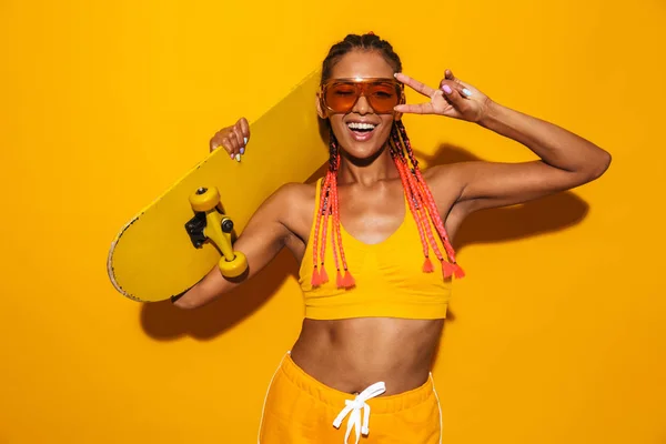 Imagen de una divertida mujer afroamericana con gafas de sol sosteniendo monopatín y haciendo gestos de señal de paz — Foto de Stock