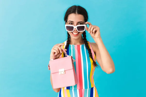 Imagen de una mujer sonriente morena con gafas de sol mirando a la cámara mientras sostiene el bolso rosa — Foto de Stock
