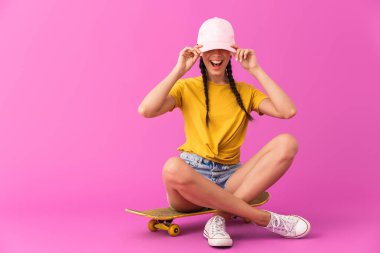 Şapka takan ve kaykay üzerinde oturan heyecanlı beyaz kadın görüntüsü