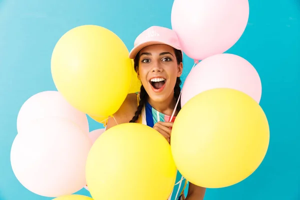 Obraz szczęśliwej kobiety zaskoczony noszenie czapki patrząc na kamerę trzymając balony powietrza — Zdjęcie stockowe